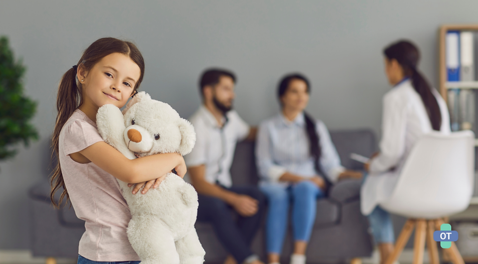 A young girl holding a teddy bear while her parents speak with an Occupational Therapist